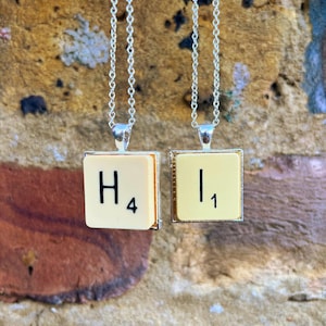 May include: Two silver chain necklaces with Scrabble tile pendants. The tiles are cream coloured with black lettering. The left tile reads "H" with the number "4" below it. The right tile reads "I" with the number "1" below it.