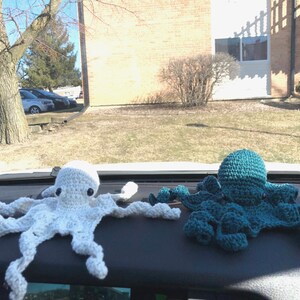 May include: Two crocheted octopus plushies, one white and one teal blue, are displayed on a car dashboard.
