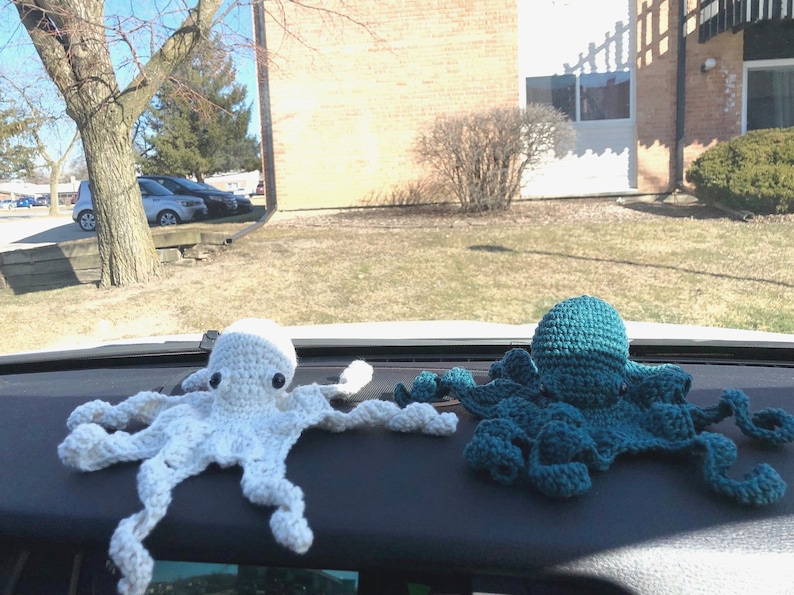 May include: Two crocheted octopus plushies, one white and one teal blue, are displayed on a car dashboard.