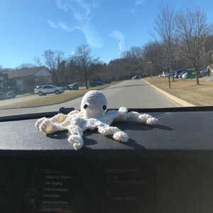 May include: A white crocheted octopus with black eyes sits on a car dashboard. The dashboard has a screen displaying the text "Settings", "Head-Up Display", "Info display", "Control display", "Time/Date", "Language/Units", "Connections", "Entertainment details", "Sia", "This Is Acting (Del...", and "Cheap Thrills".