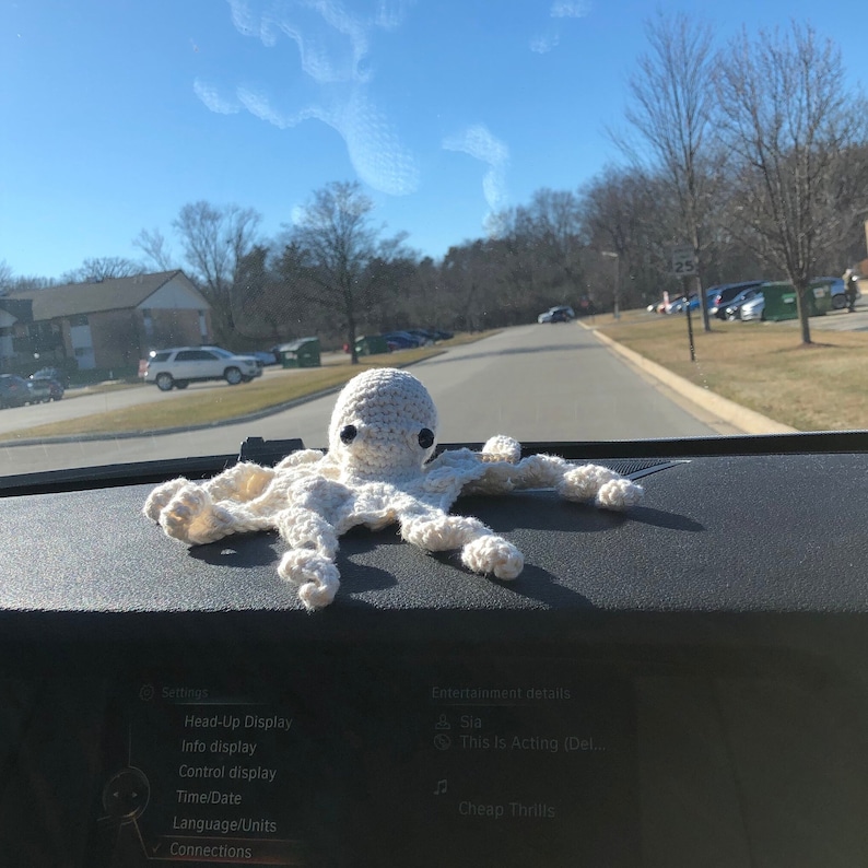 May include: A white crocheted octopus with black eyes sits on a car dashboard. The dashboard has a screen displaying the text "Settings", "Head-Up Display", "Info display", "Control display", "Time/Date", "Language/Units", "Connections", "Entertainment details", "Sia", "This Is Acting (Del...", and "Cheap Thrills".