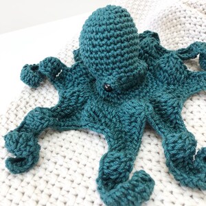 May include: A teal blue crocheted octopus with eight tentacles. The octopus is lying on a white crocheted blanket.