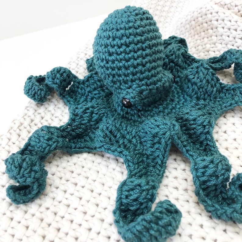May include: A teal blue crocheted octopus with eight tentacles. The octopus is lying on a white crocheted blanket.