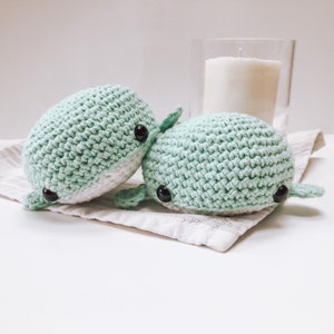 May include: Two crocheted whale plushies in a light green colour with black eyes. The plushies are sitting on a light brown fabric surface.