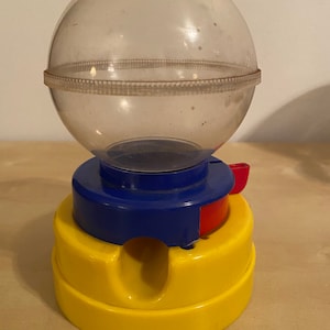 May include: A clear plastic gumball machine with a yellow base and a blue and red top. The machine has a crank on the side to dispense the gumballs.