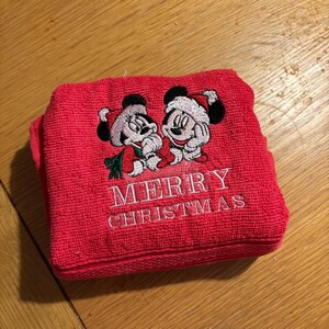 Puede incluir: Toalla roja con Mickey Mouse y Minnie Mouse bordados con gorros de Pap&aacute; Noel. La toalla dice "Feliz Navidad" en bordado blanco.