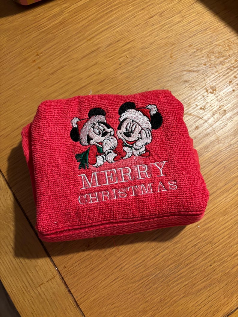 Puede incluir: Toalla roja con Mickey Mouse y Minnie Mouse bordados con gorros de Pap&aacute; Noel. La toalla dice "Feliz Navidad" en bordado blanco.