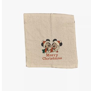 Puede incluir: Toalla de mano blanca con una imagen bordada de Mickey Mouse y Minnie Mouse con gorros de Pap&aacute; Noel. El texto "Merry Christmas" est&aacute; bordado debajo de la imagen.