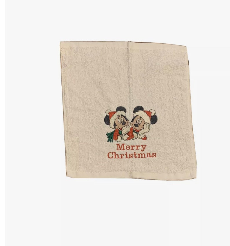 Puede incluir: Toalla de mano blanca con una imagen bordada de Mickey Mouse y Minnie Mouse con gorros de Pap&aacute; Noel. El texto "Merry Christmas" est&aacute; bordado debajo de la imagen.