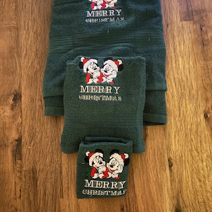 Puede incluir: Conjunto de tres toallas verdes con personajes bordados de Mickey Mouse y Minnie Mouse. Las toallas tienen el texto "Merry Christmas" bordado.