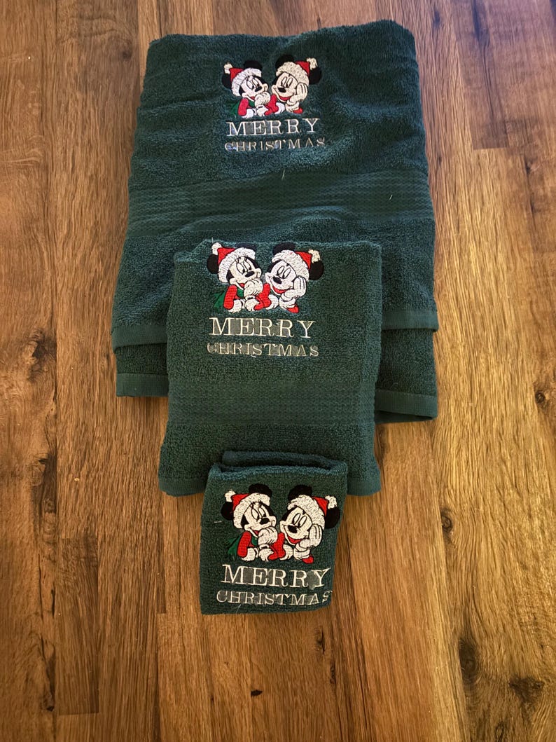 Puede incluir: Conjunto de tres toallas verdes con personajes bordados de Mickey Mouse y Minnie Mouse. Las toallas tienen el texto "Merry Christmas" bordado.