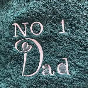 Peut inclure: Une serviette vert foncé avec le texte brodé "NO 1 Dad" en fil rose clair. Le numéro 1 et le mot "Dad" sont dans une police stylisée. La serviette a un aspect texturé et moelleux.