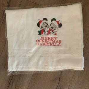 Puede incluir: Toalla blanca con texto bordado en rojo que dice "Merry Christmas Gabriella" y una imagen de dibujos animados roja y blanca de Mickey Mouse y Minnie Mouse.
