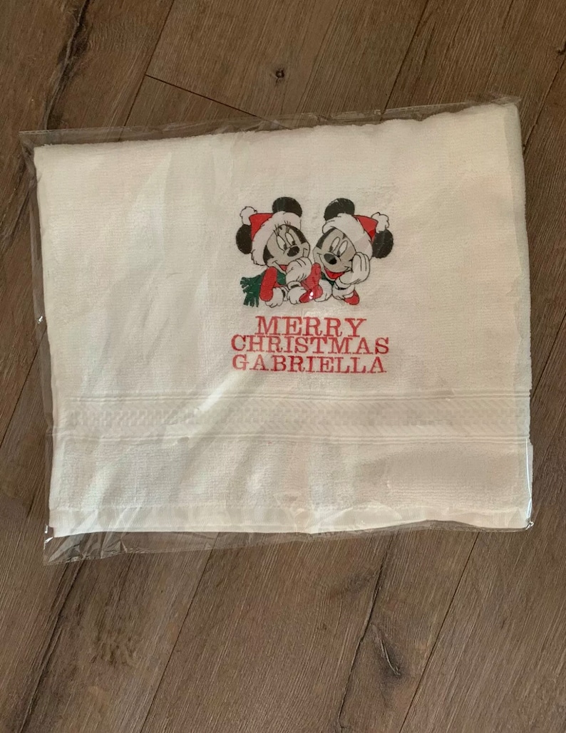 Puede incluir: Toalla blanca con texto bordado en rojo que dice "Merry Christmas Gabriella" y una imagen de dibujos animados roja y blanca de Mickey Mouse y Minnie Mouse.