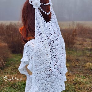 May include: A white, crocheted shawl and headpiece. The headpiece features a floral design and beaded accents. The shawl has a delicate, openwork pattern. The text "Crochetverse" is visible on the garment. The overall theme is bridal or bohemian.