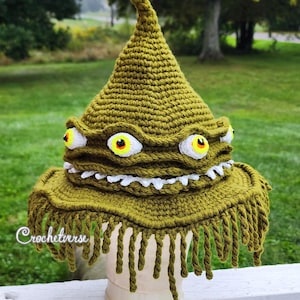 Puede incluir: Un sombrero de ganchillo puntiagudo de color verde oliva con múltiples ojos saltones de iris amarillos y esclerótica blanca, una hilera de dientes blancos dentados en el borde y largos flecos retorcidos. El texto Crochetverse es visible.