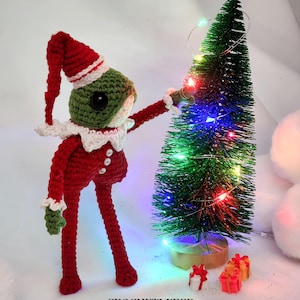 Puede incluir: Una rana de crochet verde que lleva un gorro de Papá Noel rojo y blanco y un disfraz de elfo está decorando un pequeño árbol de Navidad artificial con luces de colores. La rana está de pie sobre una superficie blanca con dos pequeños regalos envueltos.