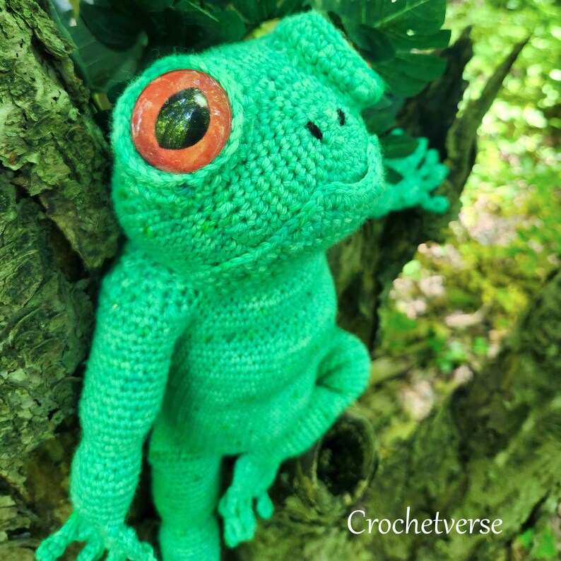 May include: A green crocheted frog with large red eyes. The frog is sitting on a tree trunk with green moss.