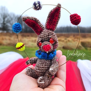 May include: A brown crocheted rabbit with red ears and a blue scarf is juggling colorful balls on a wire hoop. The rabbit has red eyes and a toothy grin. The background is a blurry image of a field and a red blanket.