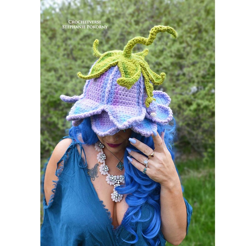 May include: A purple and blue crocheted flower hat with green crocheted leaves and stem. The hat is worn by a person with blue hair.