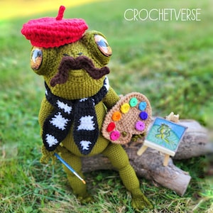 May include: A green crocheted frog wearing a red beret and a black and white scarf holds a brown paint palette with colourful paint dots. The frog is holding a blue paintbrush and is sitting on a tree branch. The image includes the text "CROCHETVERSE".