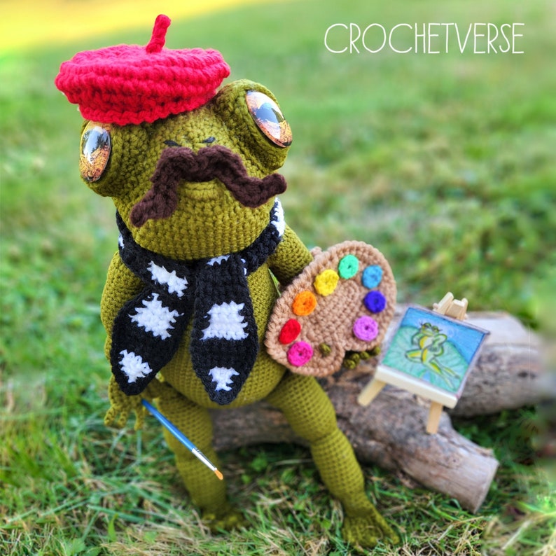 May include: A green crocheted frog wearing a red beret and a black and white scarf holds a brown paint palette with colourful paint dots. The frog is holding a blue paintbrush and is sitting on a tree branch. The image includes the text "CROCHETVERSE".