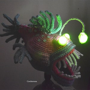 K&ouml;nnte beinhalten: Eine geh&auml;kelte Anglerfisch-Skulptur mit leuchtend gr&uuml;ner Angel und Augen. Der Fisch hat einen strukturierten K&ouml;rper in Rosa-, Lila- und Gr&uuml;nt&ouml;nen, mit wei&szlig;en Z&auml;hnen und gr&uuml;nen R&uuml;schen. Der Text "Crochetverse" ist sichtbar.