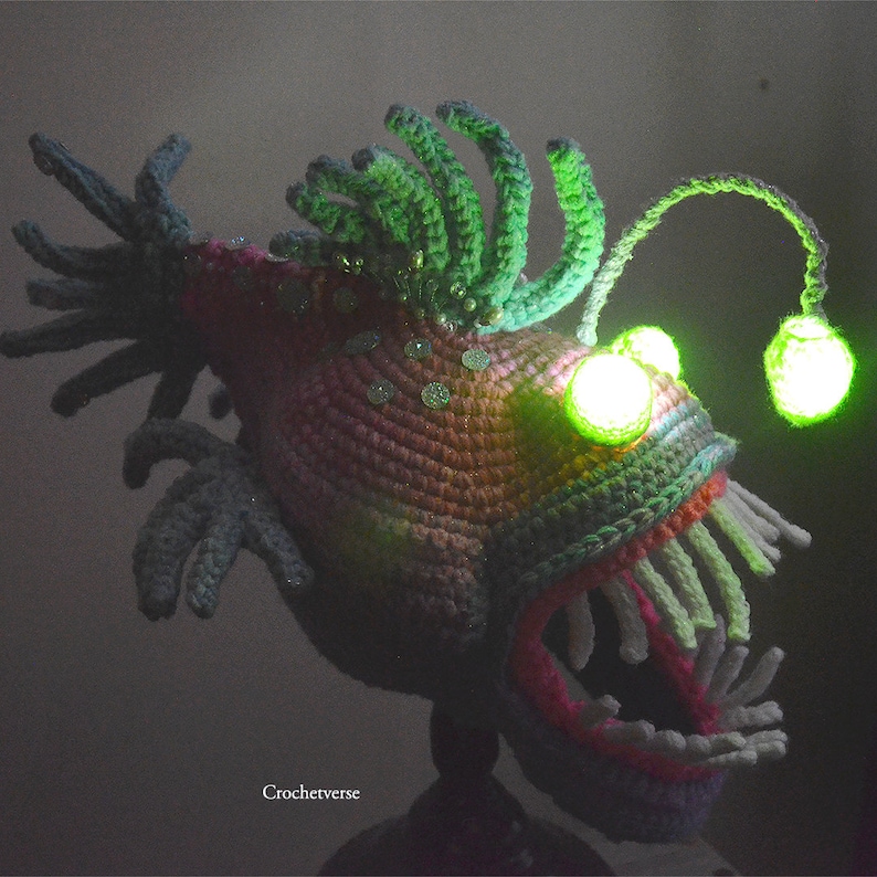 K&ouml;nnte beinhalten: Eine geh&auml;kelte Anglerfisch-Skulptur mit leuchtend gr&uuml;ner Angel und Augen. Der Fisch hat einen strukturierten K&ouml;rper in Rosa-, Lila- und Gr&uuml;nt&ouml;nen, mit wei&szlig;en Z&auml;hnen und gr&uuml;nen R&uuml;schen. Der Text "Crochetverse" ist sichtbar.