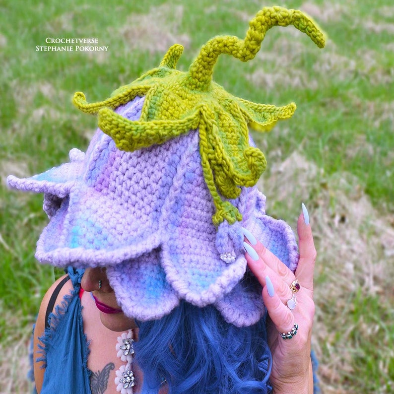 May include: A crocheted flower hat with a purple and blue base and a green stem. The hat is worn by a person with blue hair.