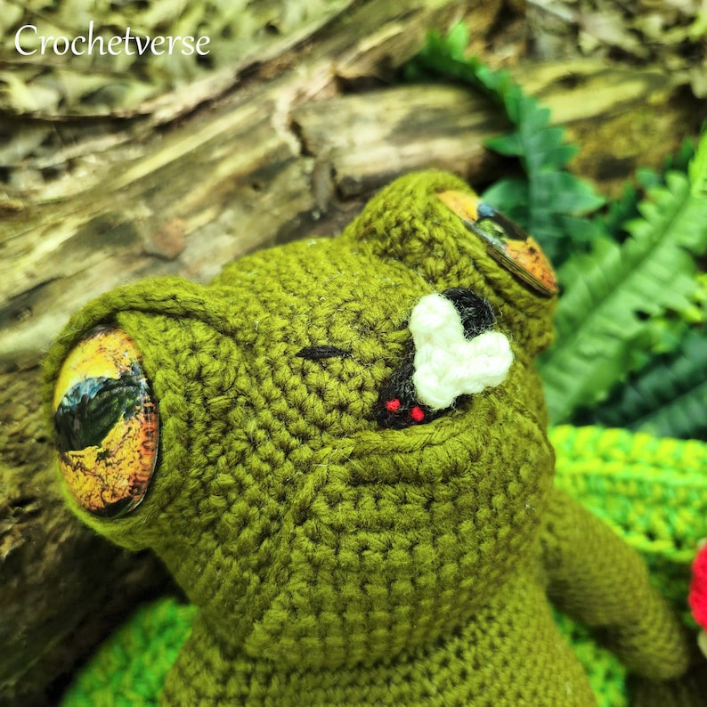 May include: Green crocheted frog with large, colourful eyes and a white crocheted nose. The frog is lying on its back with its mouth open.