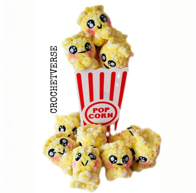 May include: A whimsical crochet popcorn design. The popcorn is yellow with pink cheeks and black eyes, spilling from a red and white striped popcorn container. The container has a "POP CORN" label. The text "CROCHETVERSE" is on the left side.