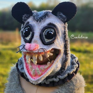 May include: A crocheted opossum balaclava featuring a white face, black ears, and large dark eyes. The mouth is open, revealing pink gums and individual off-white teeth. The text Crochetverse is visible on the right side of the head piece.