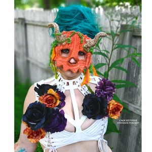 May include: A person wearing a crocheted pumpkin mask with horns and vines. The mask is orange and has a skull-like design. The person is also wearing a white top with black and purple flowers.