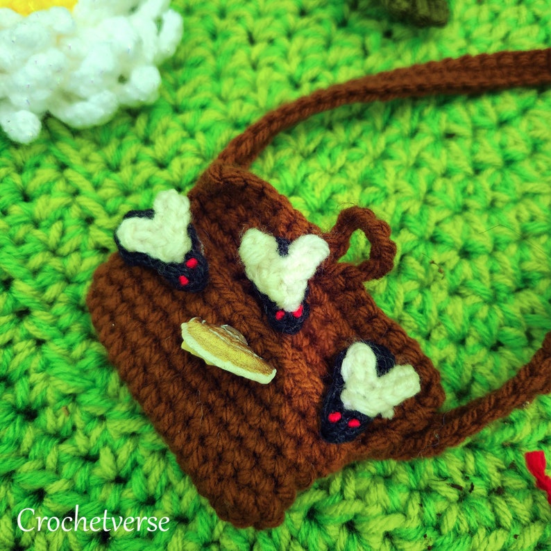 May include: A brown crocheted bag with a strap, featuring three white heart-shaped appliques with black accents. The bag is on a green crocheted surface.