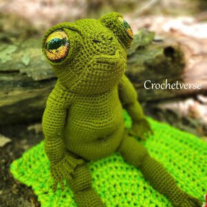 May include: A green crocheted frog with large, orange and green eyes. The frog is sitting on a green crocheted blanket. The text "Crochetverse" is visible in the background.