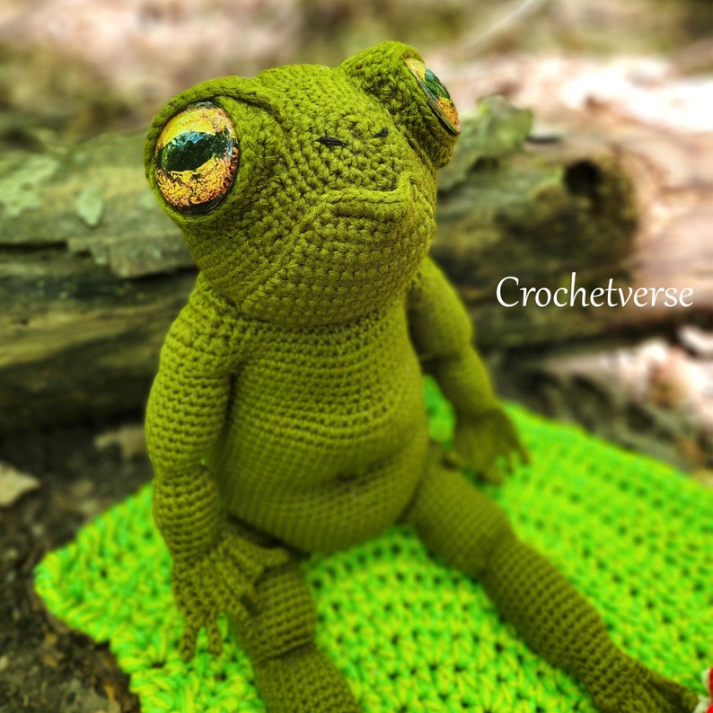May include: A green crocheted frog with large, orange and green eyes. The frog is sitting on a green crocheted blanket. The text "Crochetverse" is visible in the background.