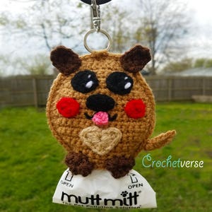 May include: A crocheted dog-shaped poop bag holder with a brown body, black nose, pink tongue, and red cheeks. The holder is attached to a silver key chain and is holding a white poop bag with the text "Mutt Mitt The Pet Pollution Solution" printed on it.
