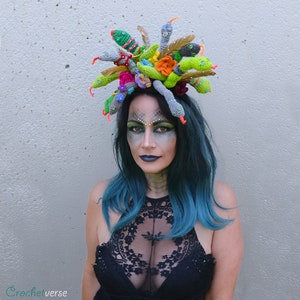 May include: A woman wearing a black lace top and a crocheted snake headpiece with colorful flowers. The headpiece is made of green, gray, and white yarn.