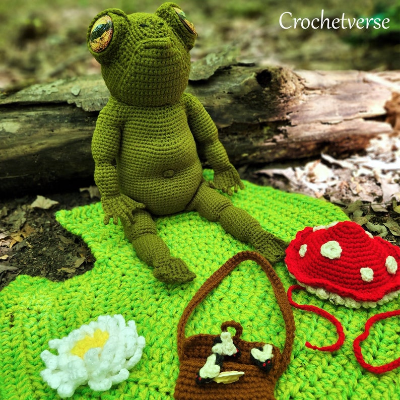 May include: A green crocheted frog sitting on a green crocheted lily pad with a red crocheted mushroom, a brown crocheted bag, and a white crocheted flower. The image is captioned "Crochetverse".