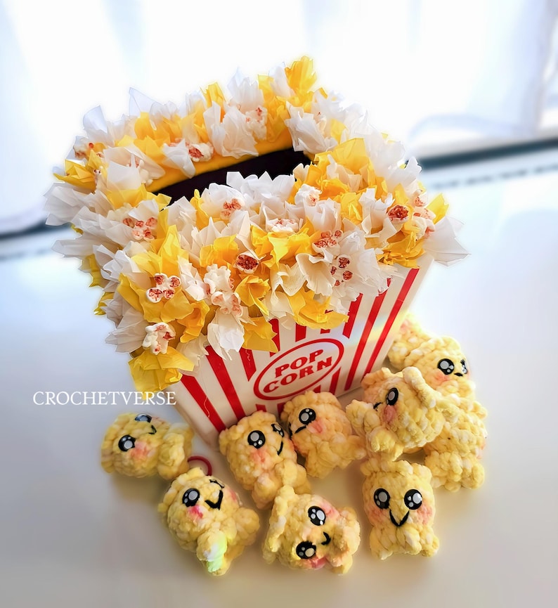 May include: A whimsical popcorn-themed display. A red and white striped popcorn container is filled with yellow and white paper flowers. Several crocheted popcorn pieces with smiling faces are scattered around the container. The text "POP CORN" is visible on the container.