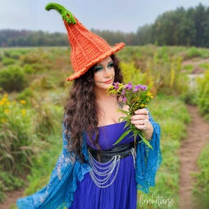 May include: An orange and green crochet witch hat with a pointed top. The hat is worn with a blue dress and a blue lace shawl. The person is holding a bouquet of purple and yellow flowers. The word "Crochetverse" is in the bottom right corner.