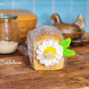 Puede incluir: Un pan envuelto adornado con una margarita de ganchillo y hojas verdes. Un tarro de cristal con una sustancia clara y una cesta de madera están en el fondo. La palabra "Crochetverse" está escrita en la superficie de madera.