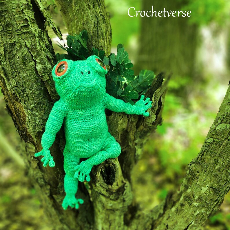 May include: A green crocheted frog with red eyes sitting on a tree branch. The frog is sitting upright with its legs crossed. The background is a blurry green forest.