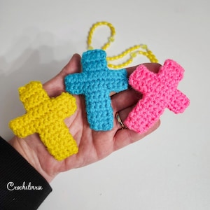 May include: Three crocheted crosses in yellow, blue, and pink are held in a hand. The crosses are connected to a yellow beaded chain. The word "Crochetverse" is written in cursive on the lower left.