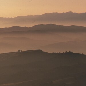 May include: A hazy, misty landscape with rolling hills and mountains in the distance. The sky is a soft pink and orange, creating a peaceful and serene atmosphere.