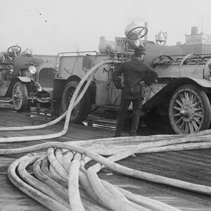May include: Two vintage fire trucks with long hoses connected to them. The trucks are parked on a paved surface. The trucks are black and white and have large wheels. The hoses are coiled on the ground.