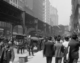 May include: A black and white photo of a busy city street with people walking and a large elevated train structure overhead. The street is lined with buildings, some with signs advertising businesses. The photo captures a moment in time in the early 20th century.