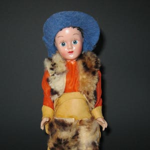 May include: A vintage doll wearing a blue hat, orange top, and fur-trimmed vest and pants. The doll has a painted face with blue eyes and is wearing yellow moccasins. The doll is a collectible item.
