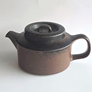 May include: A ceramic teapot featuring a two-tone design with a dark charcoal black upper section and lid, transitioning into a speckled earthy brown base. The teapot has a short spout, a curved handle, and a circular lid with a small knob handle.