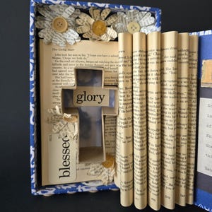 May include: A mixed media art piece featuring a cross-shaped cutout with the word "glory" and "blessed" on a book page background. The piece is framed in a blue and white patterned box, adorned with paper flowers and buttons.